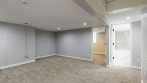 Empty finished basement with gray walls, beige carpet, recessed lights and doorways to a bedroom and a tiled bathroom